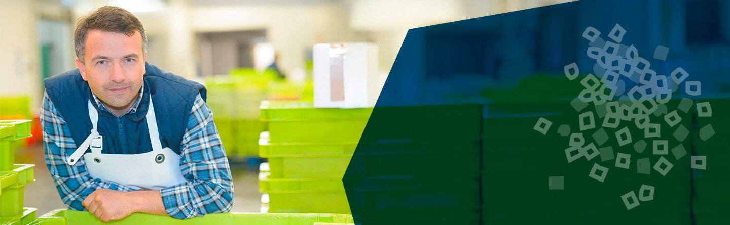 Header: In einer Produktionshalle stützt sich ein Ausbilder mit blauem Overall und einer weißen Schürze mit den Armen auf einem Tisch ab und schaut in die Kamera. Hinter und neben ihm sind grüne Kunststoffboxen zu sehen.