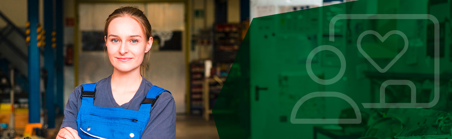 Header: Eine Handwerkerin steht, in die Kamera lächelnd, in einer Werkstatt. 