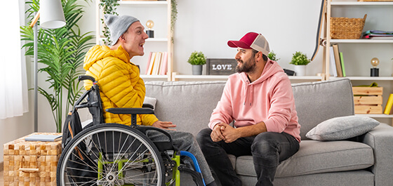 Kachelbild: Eine Betreuungsperson sitzt auf einem Sofa und unterhält sich mit einer Person im Rollstuhl