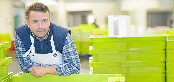 Kachelbild: In einer Produktionshalle stützt sich ein Ausbilder mit blauem Overall und einer weißen Schürze mit den Armen auf einem Tisch ab und schaut in die Kamera. Hinter und neben ihm sind grüne Kunststoffboxen zu sehen.