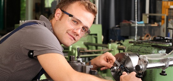 Kachelbild: Ein Maschinenbauer lächelt in die Kamera und ist mit einer Schutzbrille bekleidet über eine Werkbank gebeugt 