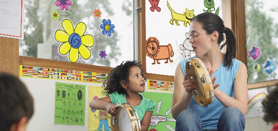 Kachelbild: Eine Erzieherin spielt in einem Kindergarten einem Kind etwas an einem Tamburin vor.