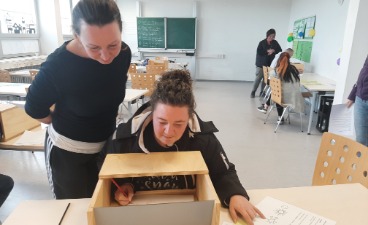 Eine Schülerin sitzt vor einer Holzkiste und bearbeitet eine Station des Demenzparcours. Eine weitere Schülerin steht hinter ihr und blick über ihre Schulter.