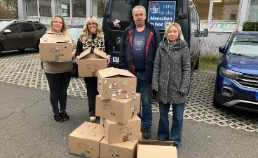 Auf dem Bild sind zwei Schülerinnen und zwei Mitarbeitende von der Organisation Space-Eye e.V. zu sehen. Sie stehen auf dem Parkplatz hinter einem Stapel gepackter Kartons. Die Schülerinnen haben jeweils ein Paket in der Hand um es den beiden Mitarbeitenden zu geben.