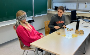 Zwei Schülerinnen sitzen an einem Tisch. Eine Schülerin hat sich die Augen verbunden. Sie muss erraten, welches Musikinstrument die andere Schülerin ihr vorspielt.