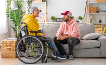 Das Bild zeigt zwei Männer, die sich ansehen und anlächeln. Der eine Mann sitzt in einem Rollstuhl vor dem Sofa, auf dem der andere sitzt.