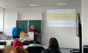 Die beiden Vertreter:innen des Berufsverbands stehen vor der Tafel und sprechen zur Gruppe, die vor ihnen sitzt. Der Beamer projiziert die Präsentation des Berufsverbands an die Wand neben der Tafel.