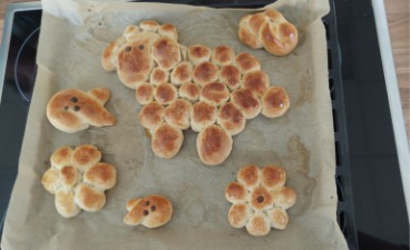 Auf einem, mit Backpapier ausgelegten Blech, liegen gebackene Hefeteig-Stück: ein Schaf, drei Hasen und zwei Blumen.