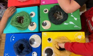 Auf den Bild sind sechs in unterschiedlich angemalte Boxen zu sehen, die mit unterschiedlich großen Augen wie Monster aussehen. Vier Kinder stecken gerade ihre Hände in die Öffnung der Kiste, die den Mund des Monsters darstellt.