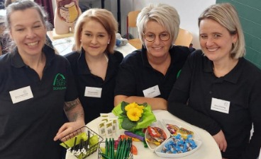Vier Frauen in schwarzem Poloshirt und mit Namensschild stehen um einen Stehtisch herum. Auf dem Tisch liegen Flyer, bfz-Kugelschreiber und Bonbons.